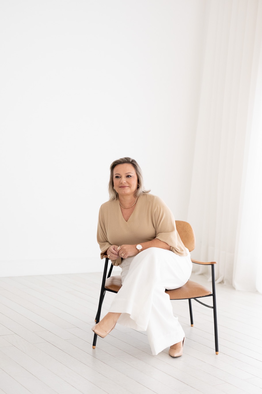 Fabiana Carvalho sentada em uma poltrona de couro caramelo, em ambiente claro, com blusa bege e calça branca.