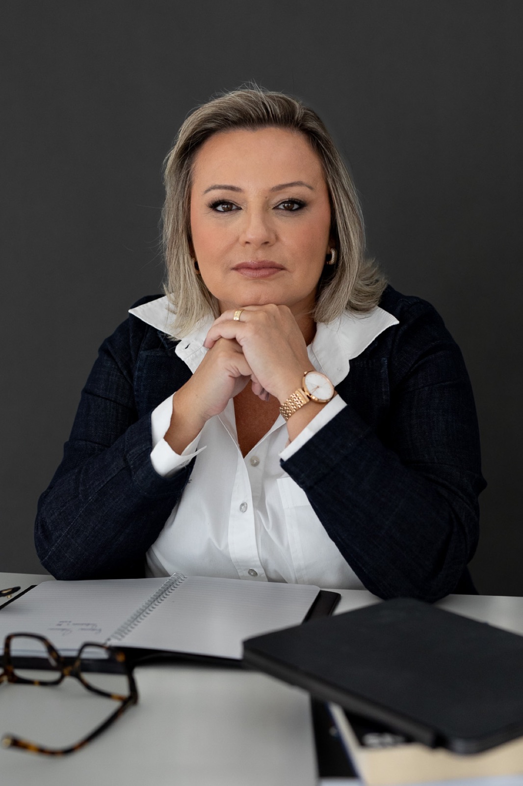 Fabiana Carvalho à mesa de trabalho, de blazer marinho e camisa branca, sorrindo com as mãos apoiadas no queixo.